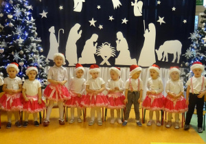 Grupa dzieci śpiewa piosenkę, w tle dekoracja świąteczna.